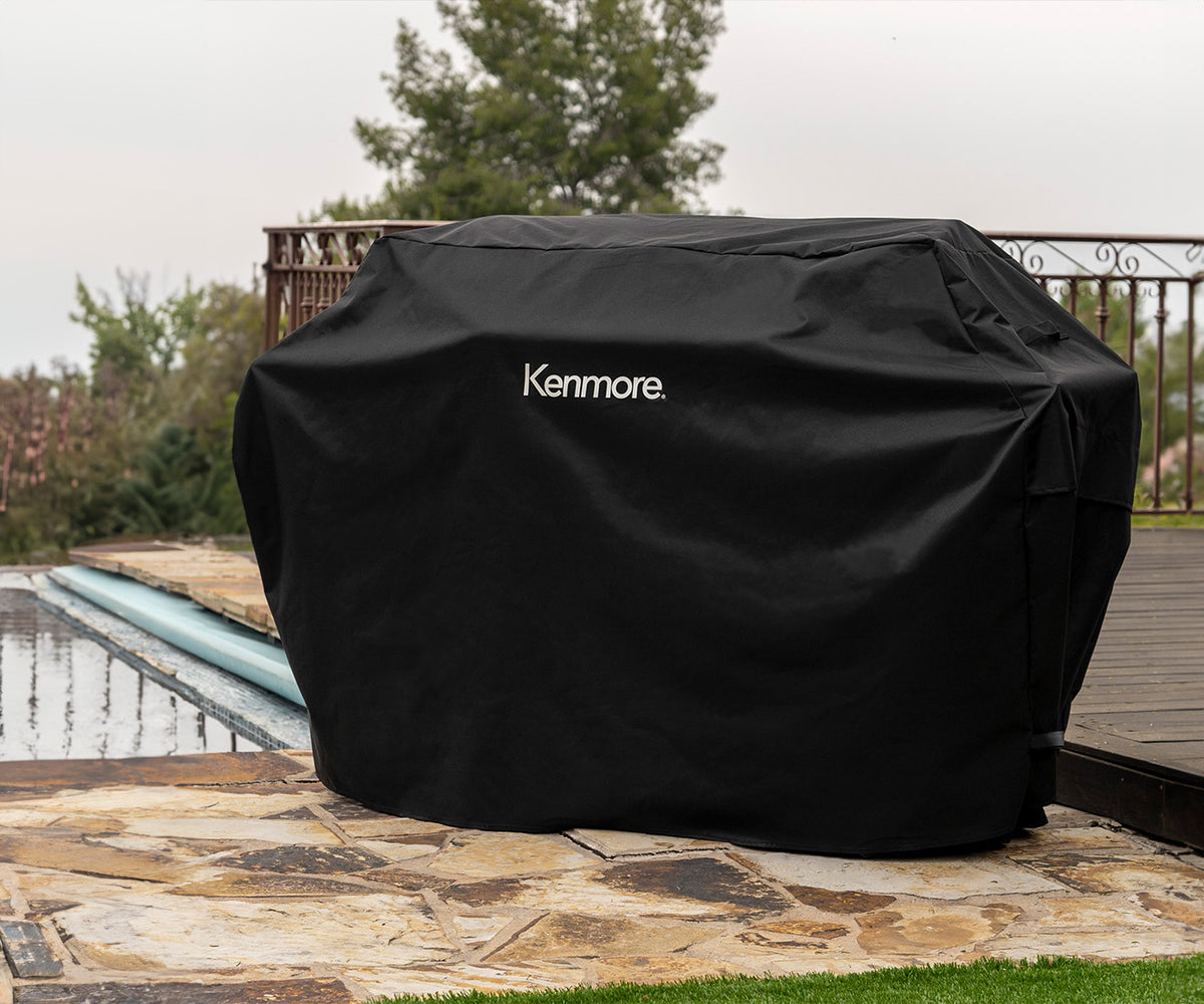 Kenmore 56-inch grill cover, black, over a grill on a stone patio beside a pool, trees and railing behind